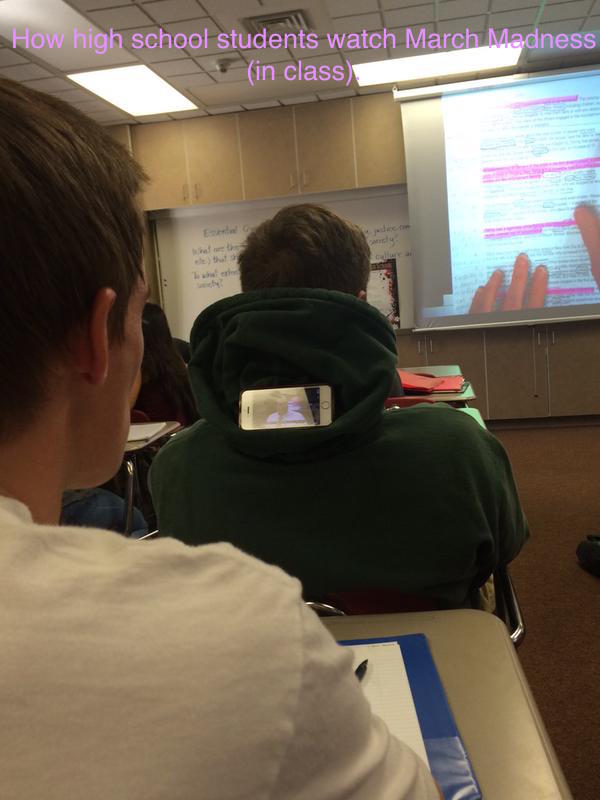 Photo of a student in class watching NCAA basketball on his iPhone, which is tucked into the hood of the student sitting in front of him.